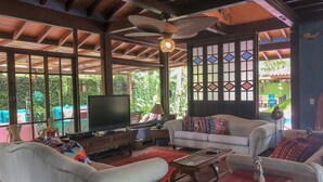 A spacious living area with a television, comfortable seating, and large windows offering a view of the outdoors. - House 50m from the beach in a unique gated community, total privacy, Camburizinho.  (Sertão do Cambury)