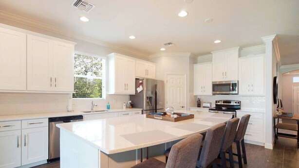 A modern kitchen with white cabinetry, a central island with bar stools, stainless steel appliances, and a view of greenery outside. - 3BR villa in gated community w/tons of amenities! Villa Eleadora - SWFL Rentals (Cape Coral)