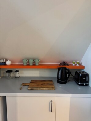 A kitchen with a wooden cutting board, a kettle, a toaster, and a shelf with cups and a teapot.