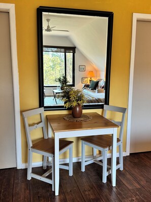 A room with a dining table, chairs, a potted plant, and a large mirror reflecting a bedroom with a bed and a ceiling fan.