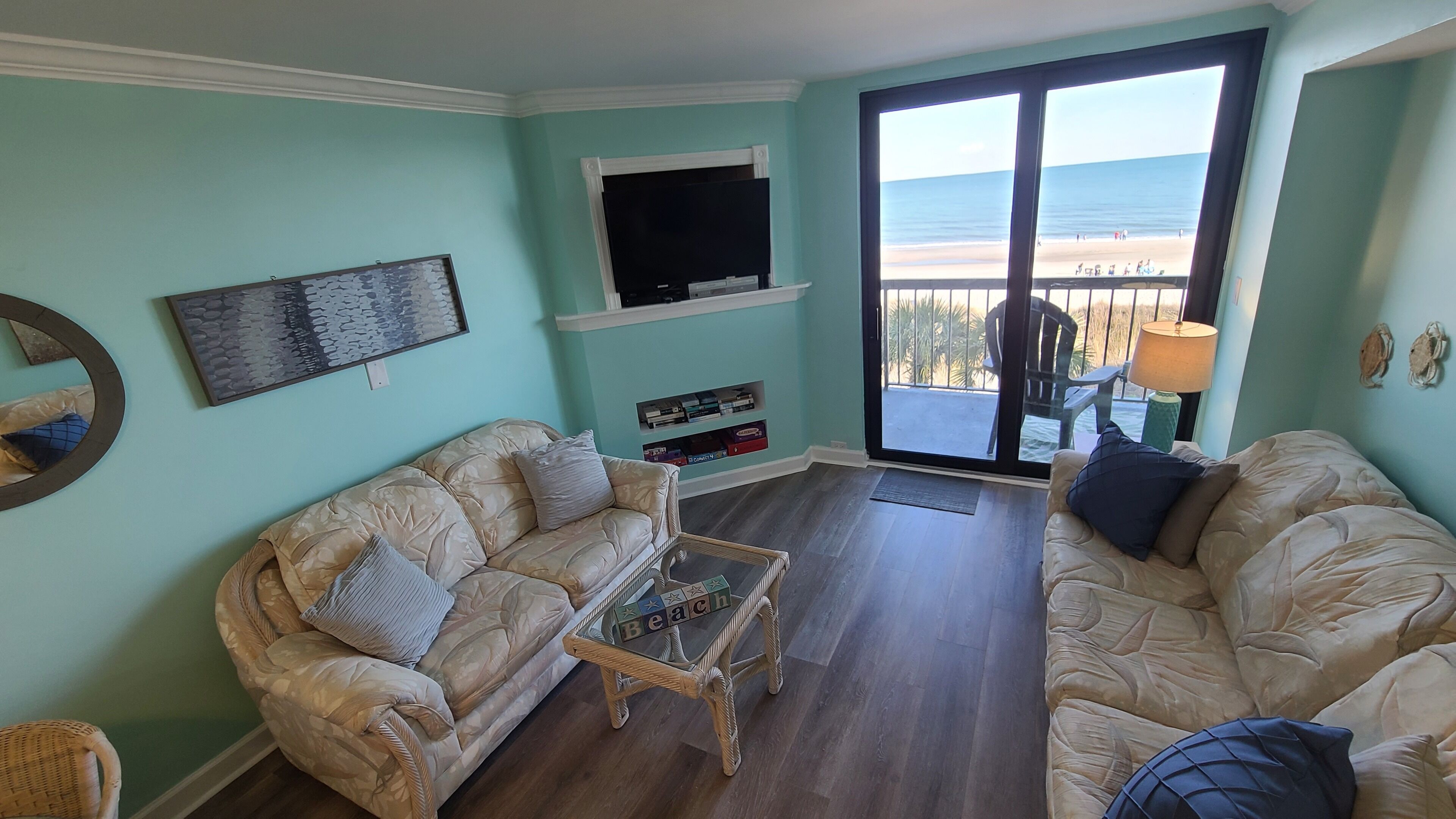 Un salon avec deux canapés beige, une table basse, un téléviseur à écran plat et une bibliothèque. Une grande fenêtre offre une vue sur la plage.