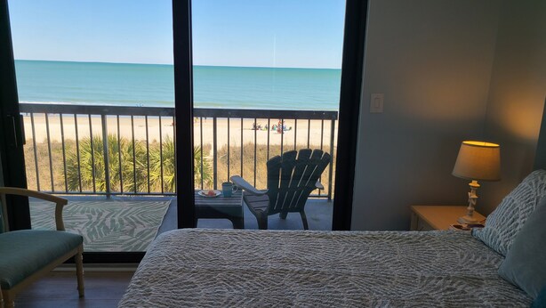 Una camera d'albergo con un letto, una sedia, un tavolino con una tazza e vista sulla spiaggia e sull'oceano da un balcone con ringhiera.