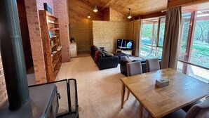 A cozy room with a brick wall, wooden ceiling, a television, a sofa, a dining table, and a large window with a view of greenery. - Grampians Private Bushland Retreat (Halls Gap)