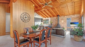 A spacious dining area with a wooden table and chairs, a large wall clock, and a kitchen with a brick wall and a chimney. - Beachcombers on Collingwood Beach (Vincentia)