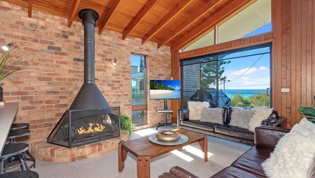 A cozy living room with a brick fireplace, leather sofa, and a large window overlooking the ocean. - Beachcombers on Collingwood Beach (Vincentia)