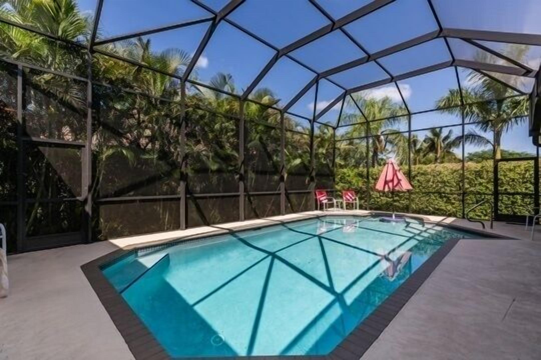 Una piscina con un cerramiento de vidrio geométrico, rodeada de palmeras y un cielo despejado.