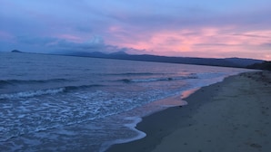 Une plage où les vagues viennent doucement lécher le rivage, un ciel dégagé et des montagnes lointaines.