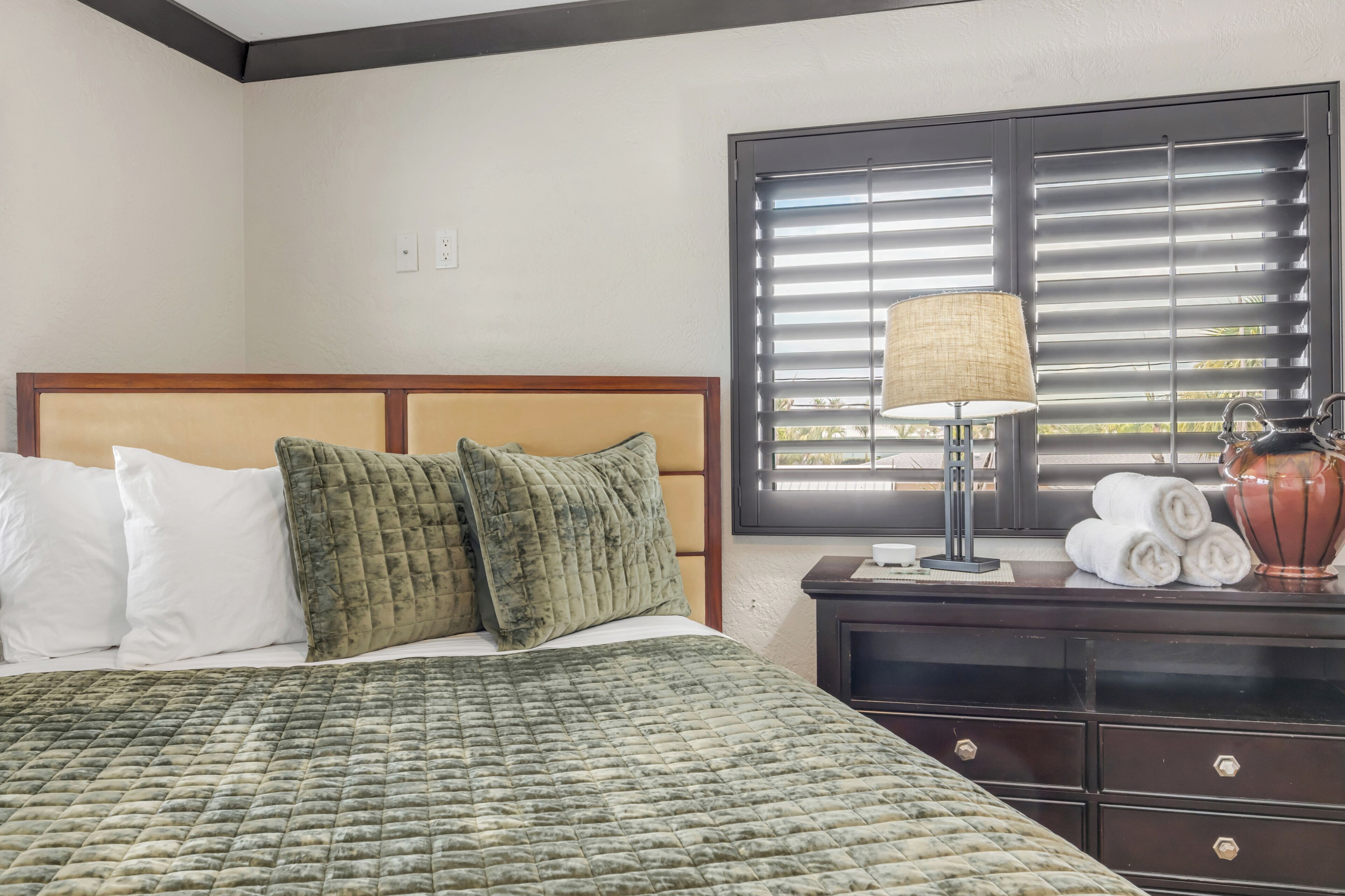 Una habitación de hotel con cama, mesita de noche, lámpara y ventana con persianas.