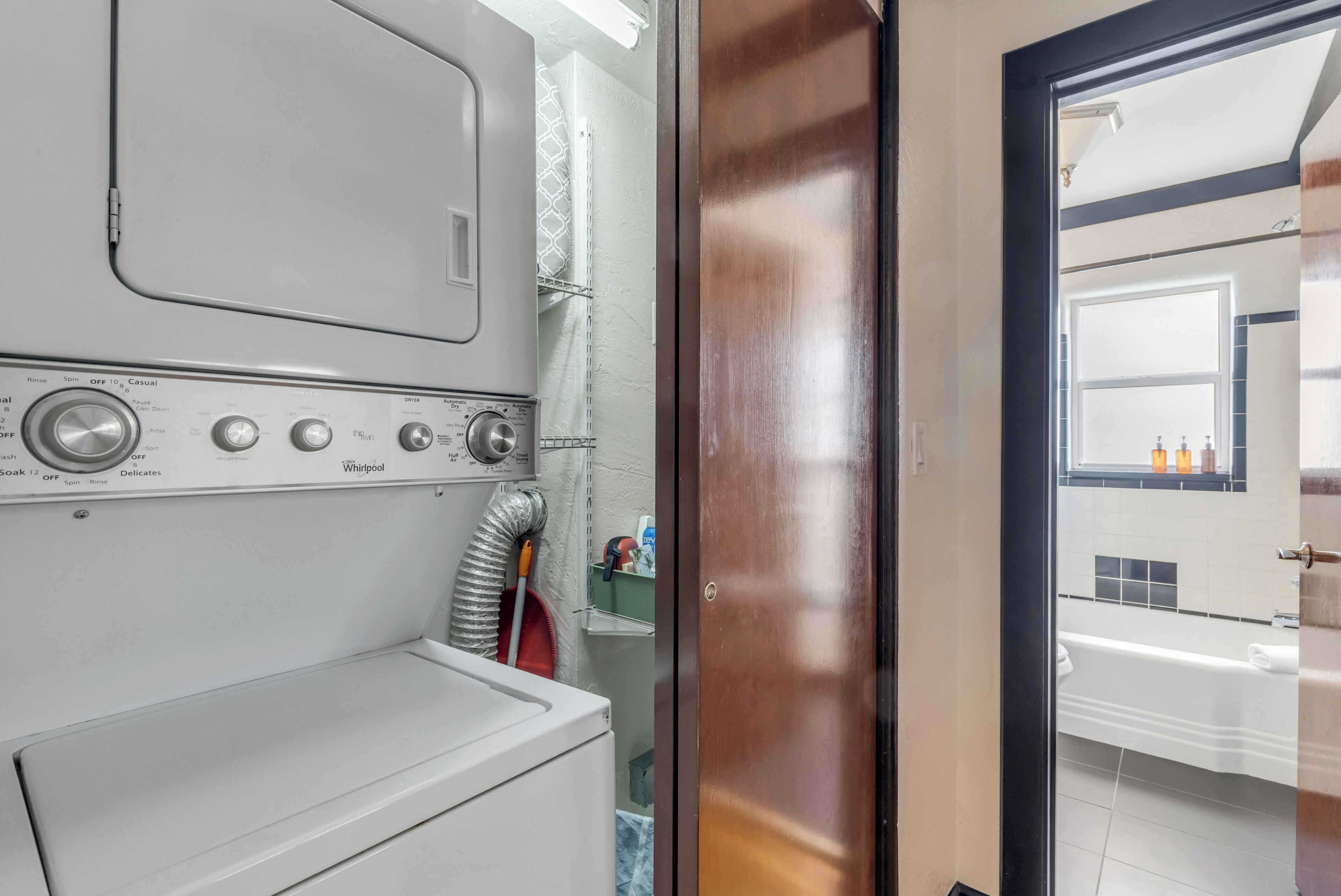 A laundry room with a front-loading washer and dryer, a vent hose, and a door leading to a bathroom with a sink and mirror.
