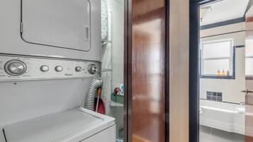 A laundry room with a front-loading washer and dryer, a vent hose, and a door leading to a bathroom with a sink and mirror.