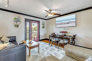 A living room with a sofa, a coffee table, a dining table with chairs, a ceiling fan, and a large TV screen displaying a beach scene.