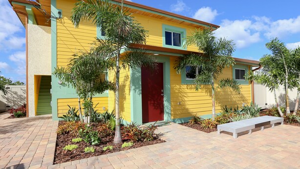 Une maison jaune avec une porte rouge, entourée de plantes et bordée d’un chemin pavé.
