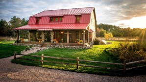 Un bâtiment de style rustique, évoquant une grange, avec un toit en métal rouge, une véranda agrémentée de plantes en pot et une allée gravillonnée.