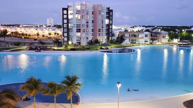 Une grande piscine à bordures courbes, entourée de palmiers et bordée d’un immeuble doté de nombreux balcons.