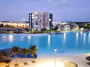 A large swimming pool with a curved edge, surrounded by palm trees and a building with multiple balconies.
