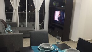 Un coin repas avec une table dressée pour quatre personnes, un canapé, un téléviseur et une fenêtre avec des rideaux.