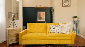 Ein gelbes Sofa mit gemusterten Kissen, ein kleiner Tisch mit einer Pflanze und ein gerahmtes Kunstwerk an der Wand.