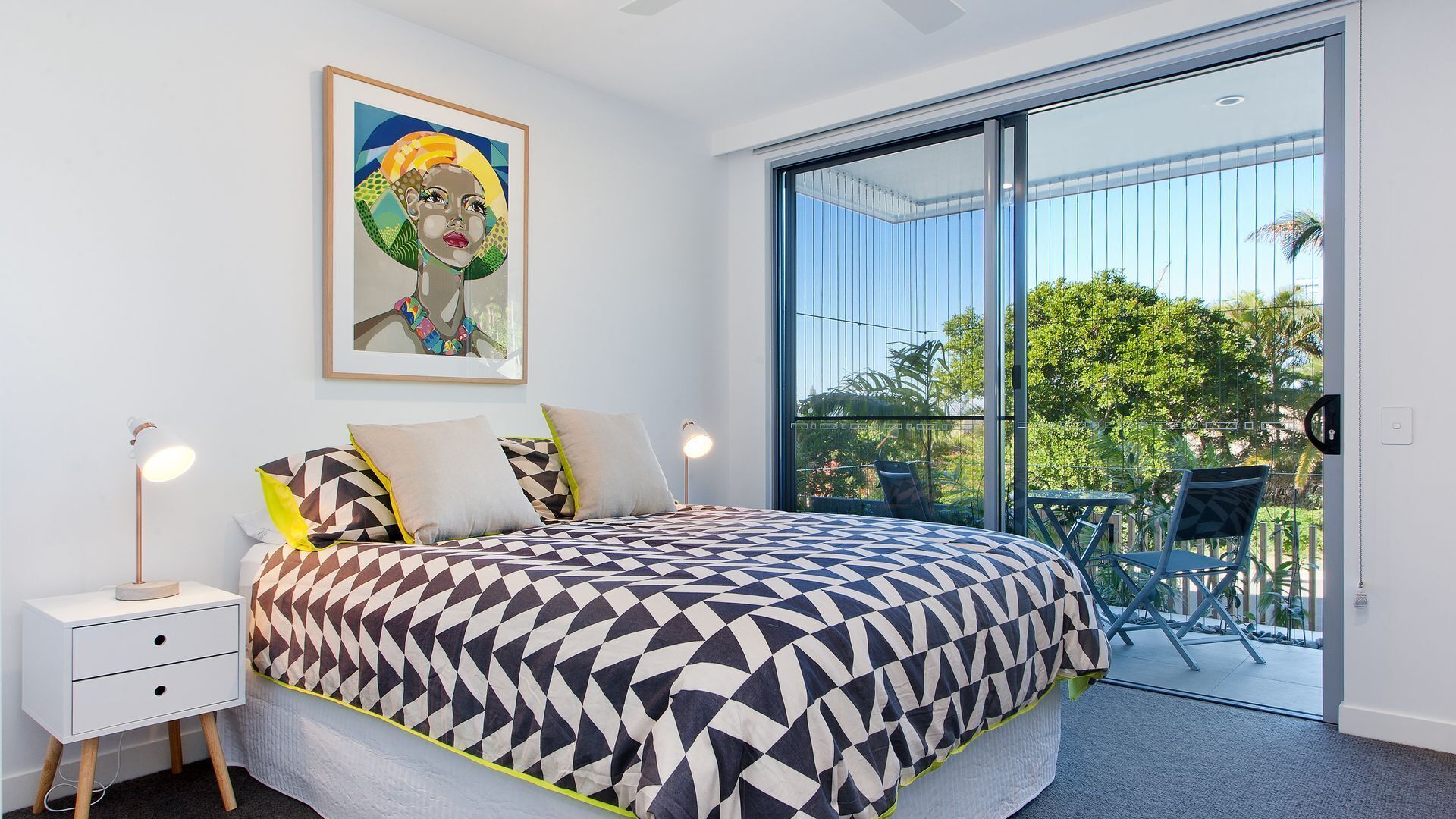 A bedroom with a large bed, a nightstand, a framed artwork, and a sliding glass door leading to an outdoor area with a chair.