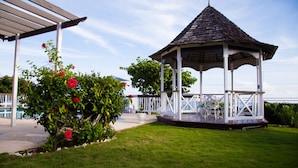 Una pérgola con techo de madera y barandillas blancas, rodeada de un césped cuidado y arbustos florecientes.