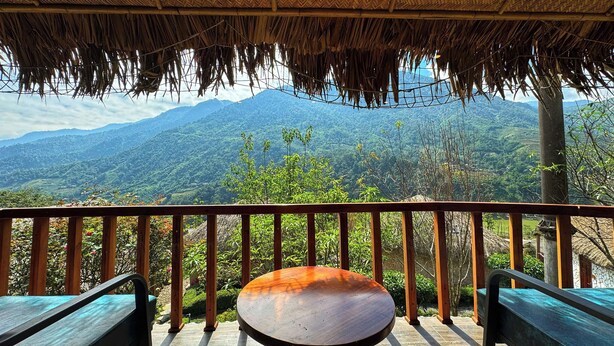 Una terraza de madera con techo de paja, una mesa redonda y dos bancos, con vistas a un paisaje montañoso.