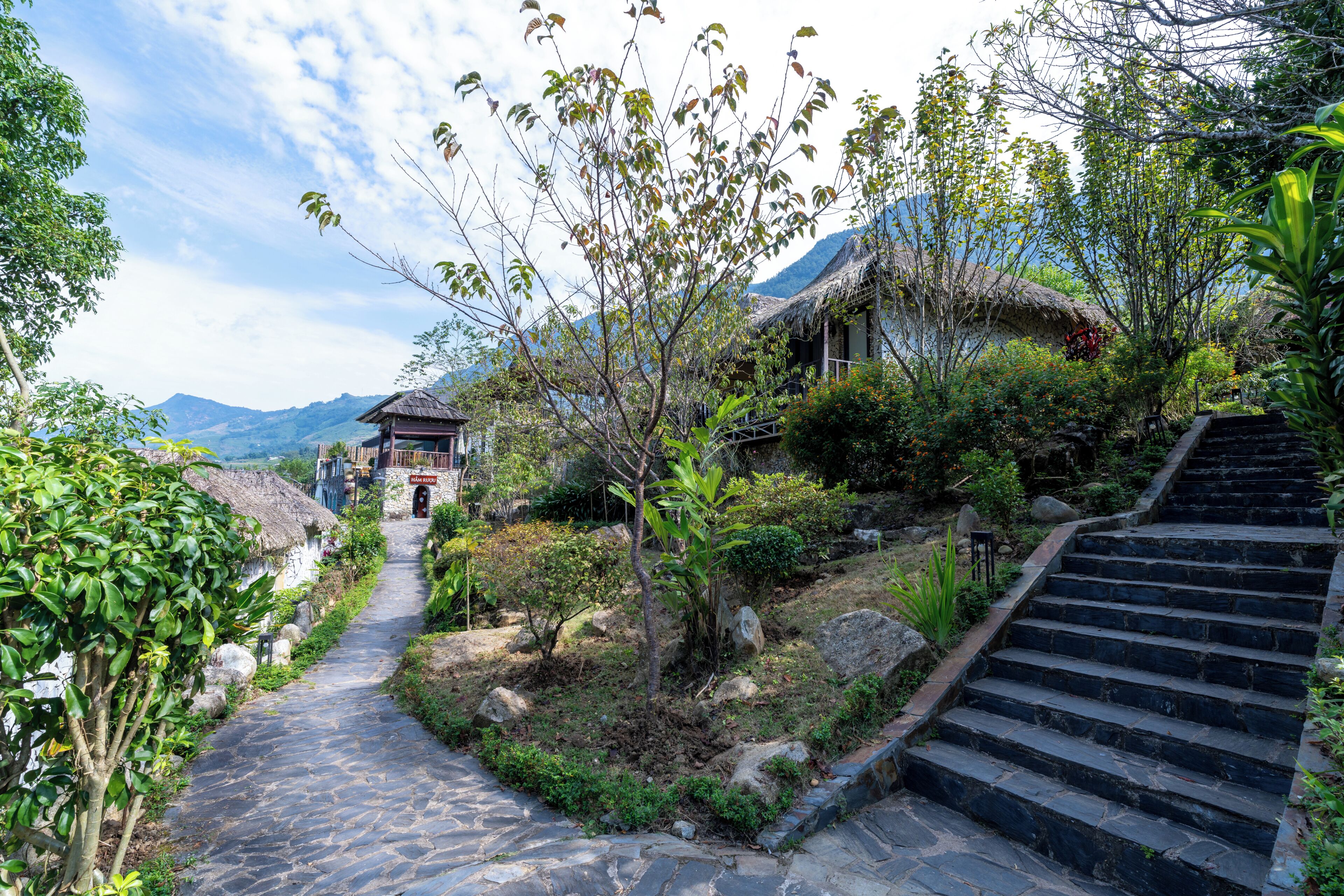 A stone pathway leading to a traditional thatched-roof building with a sign reading 'HÀM RƯU'.