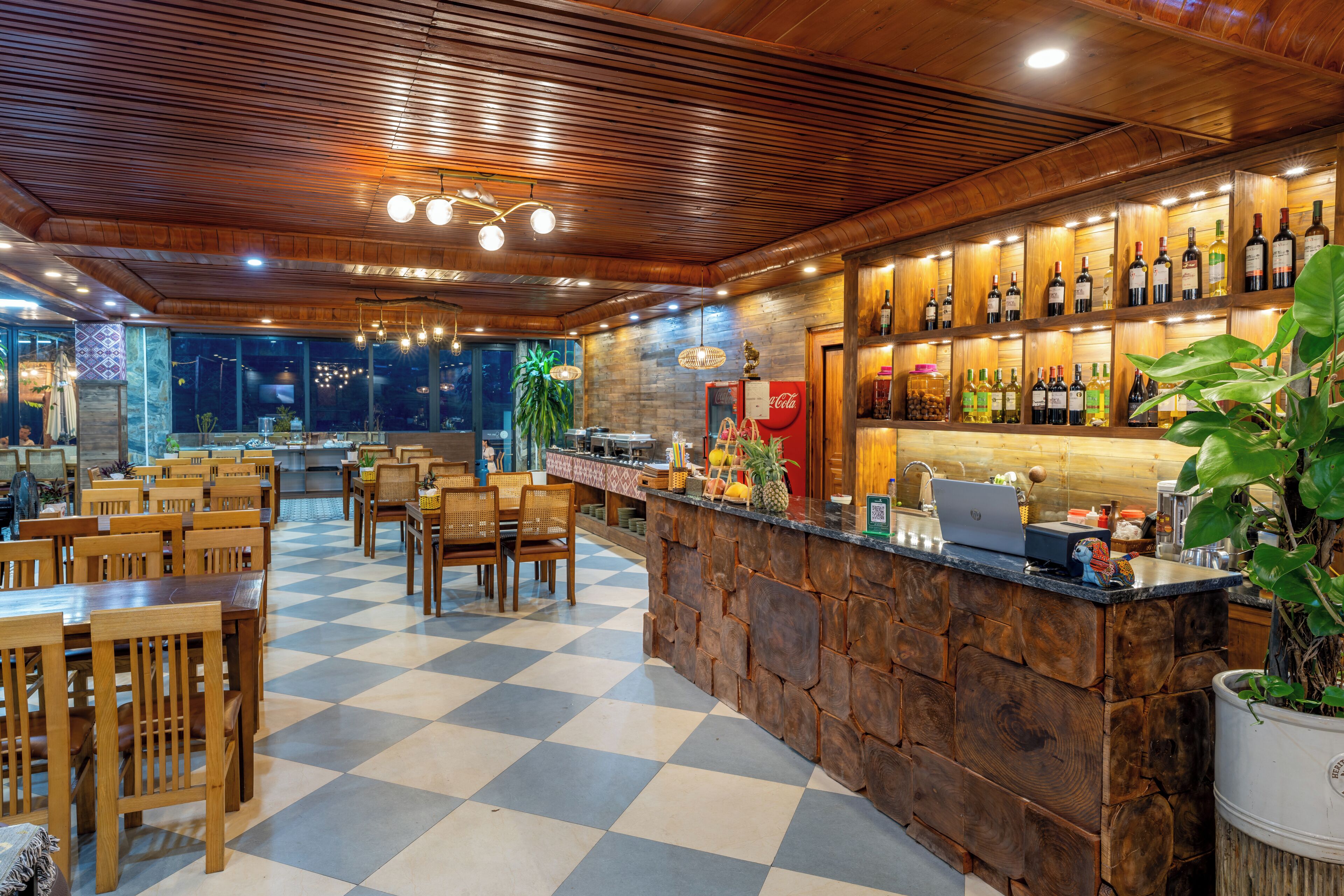 Un restaurante con piso a cuadros, mesas y sillas de madera, una zona de bar con botellas y un ordenador portátil sobre la barra.