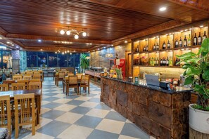 Un restaurante con piso a cuadros, mesas y sillas de madera, una zona de bar con botellas y un ordenador portátil sobre la barra.