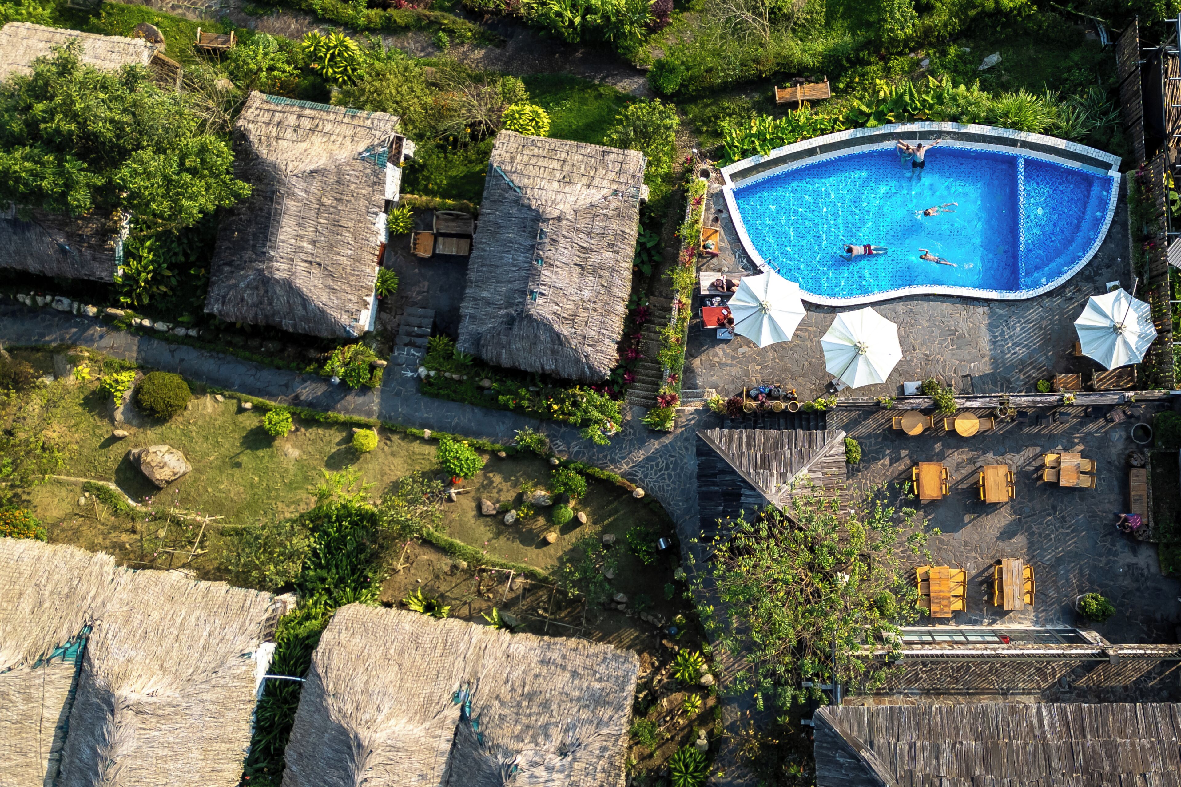 An aerial view of a resort with a swimming pool, thatched-roof structures, and outdoor seating areas.