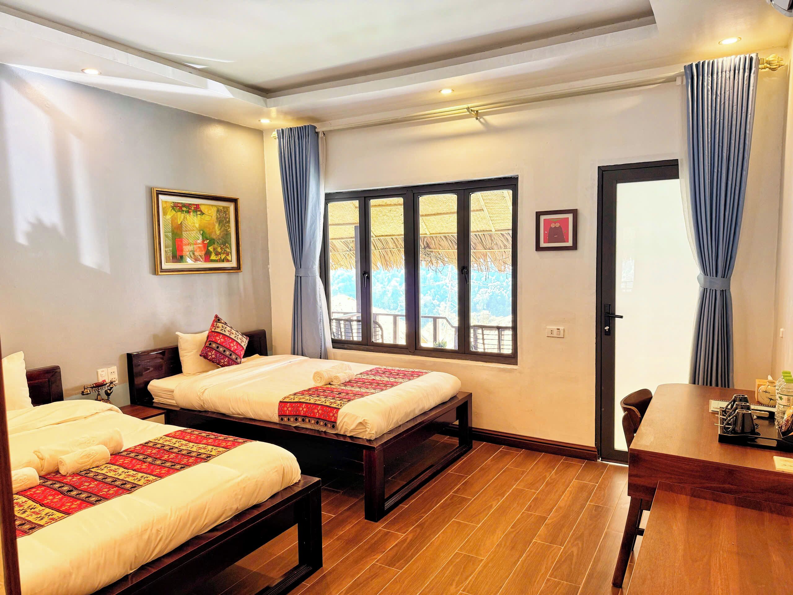 A hotel room with two beds, a wooden floor, a dining table, and a large window with curtains.