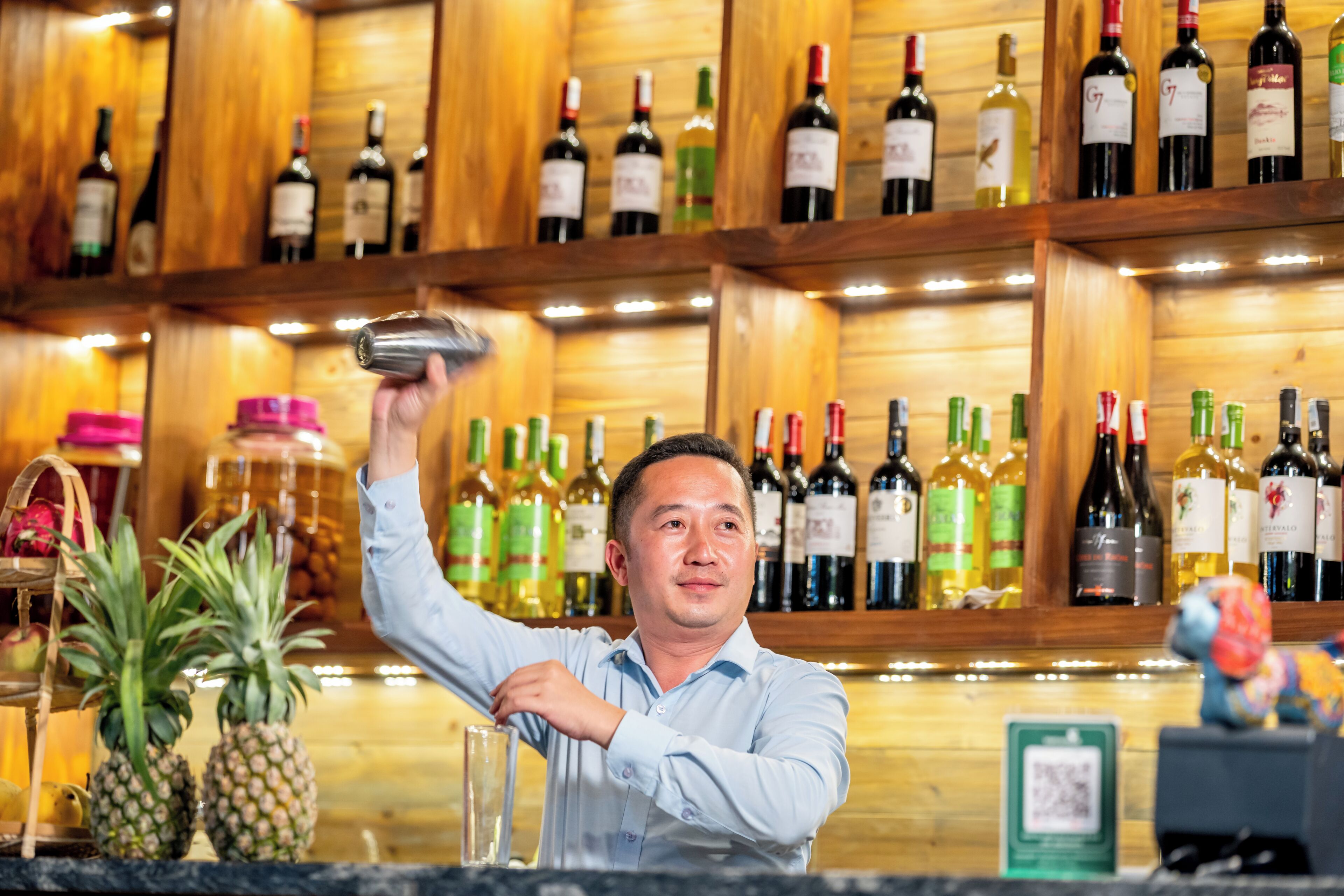 A bar setting with a person pouring a drink, shelves of wine bottles, and a pineapple on the counter.