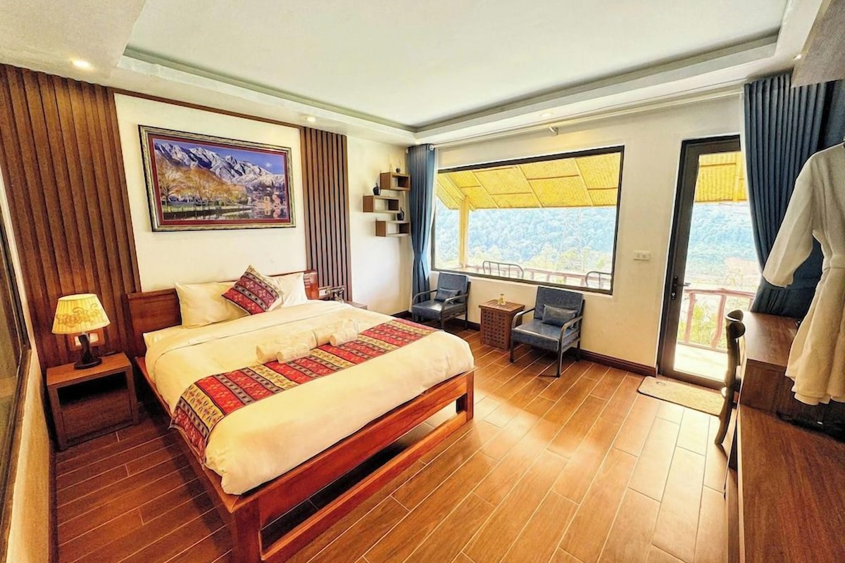 Una habitación de hotel con una cama grande, un escritorio, una silla, un ventanal con vistas y un cuadro en la pared.