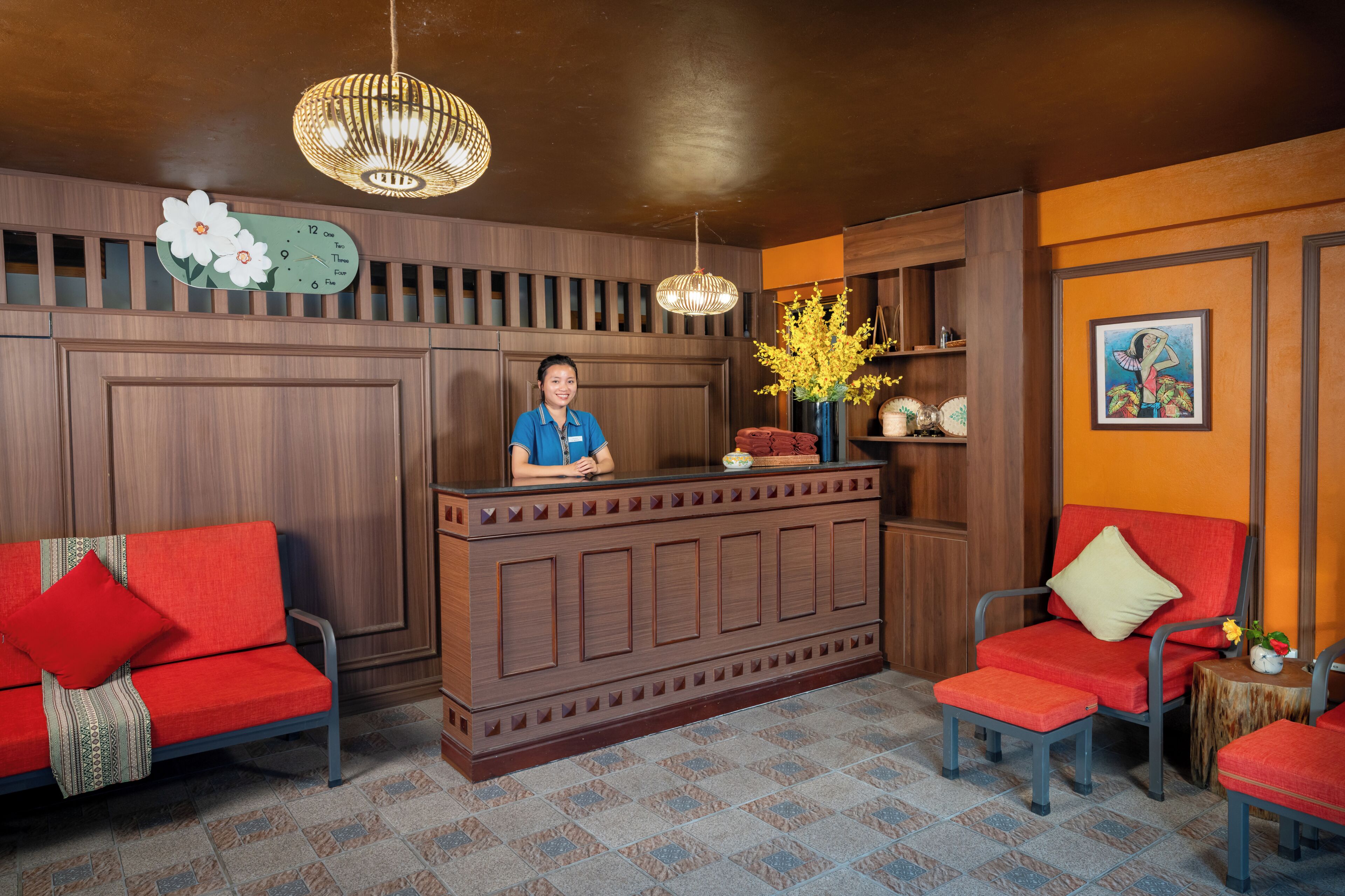 A reception area with a wooden reception desk, two red upholstered chairs, a clock, and a vase with yellow flowers.