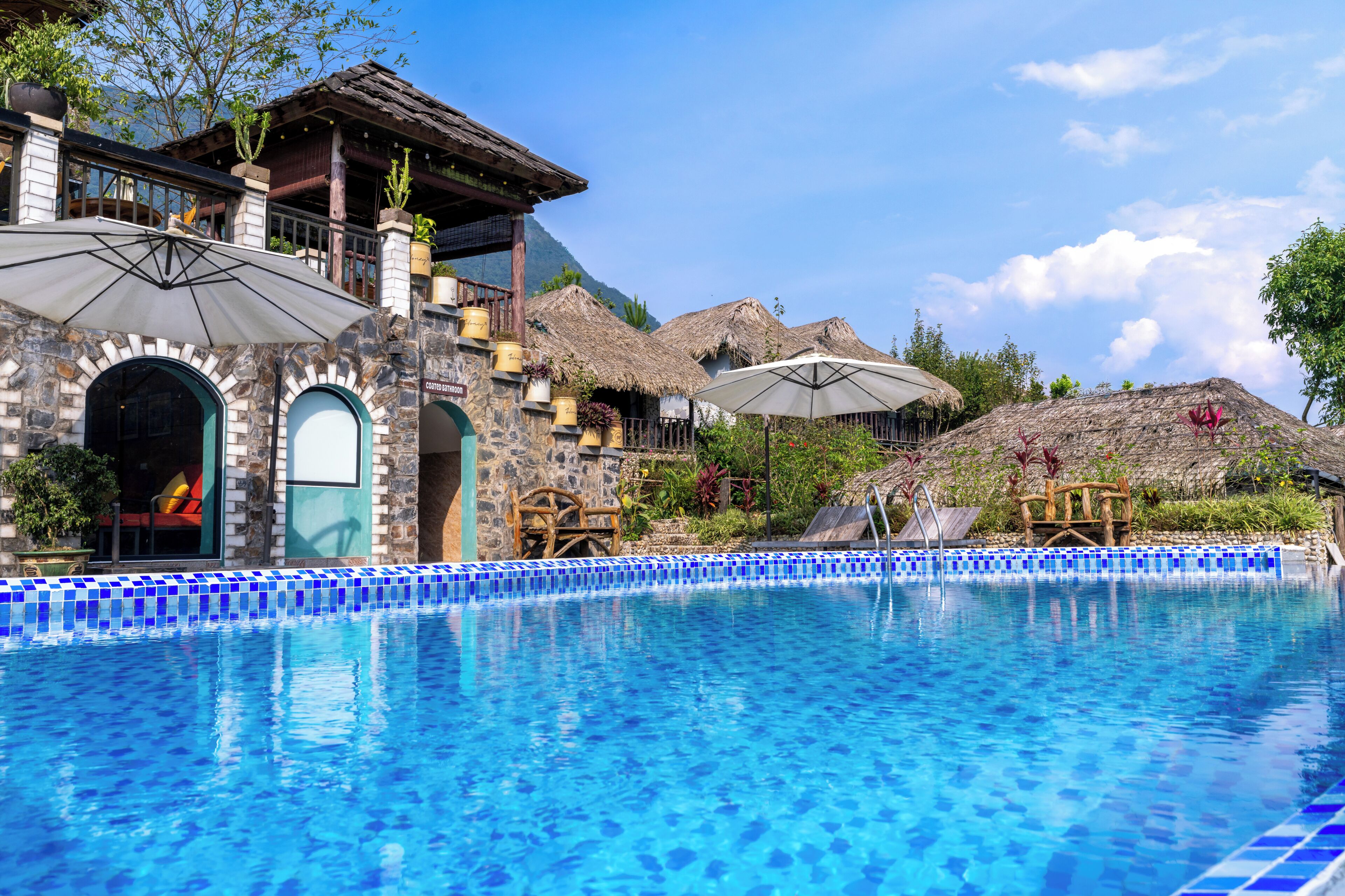 Una piscina con mosaicos azules, un baño cubierto y edificios con techos de paja.