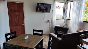 Et værelse med et træbord, sorte stole, en brun sofa, et tv monteret på væggen og et vindue med gardiner.
