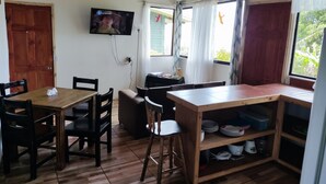 Et værelse med et træbord, stole, et fjernsyn ophængt på væggen og et vindue med gardiner.