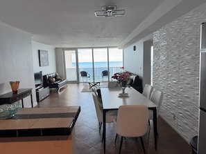 A modern living room with a dining area, a sofa, a TV, and a view of the sea.