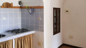Une cuisine équipée d’une cuisinière à gaz, d’un évier et d’un plan de travail recouvert de carreaux. On trouve une étagère en bois au-dessus de la cuisinière et une fenêtre sur le mur de droite.