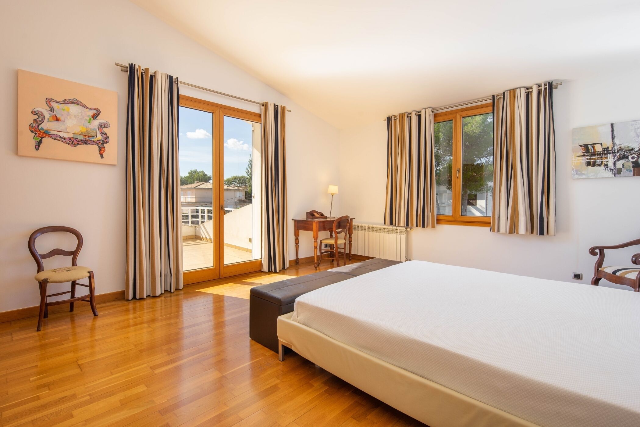 A bedroom with a large bed, wooden floor, a desk with a chair, and a view through the glass door.
