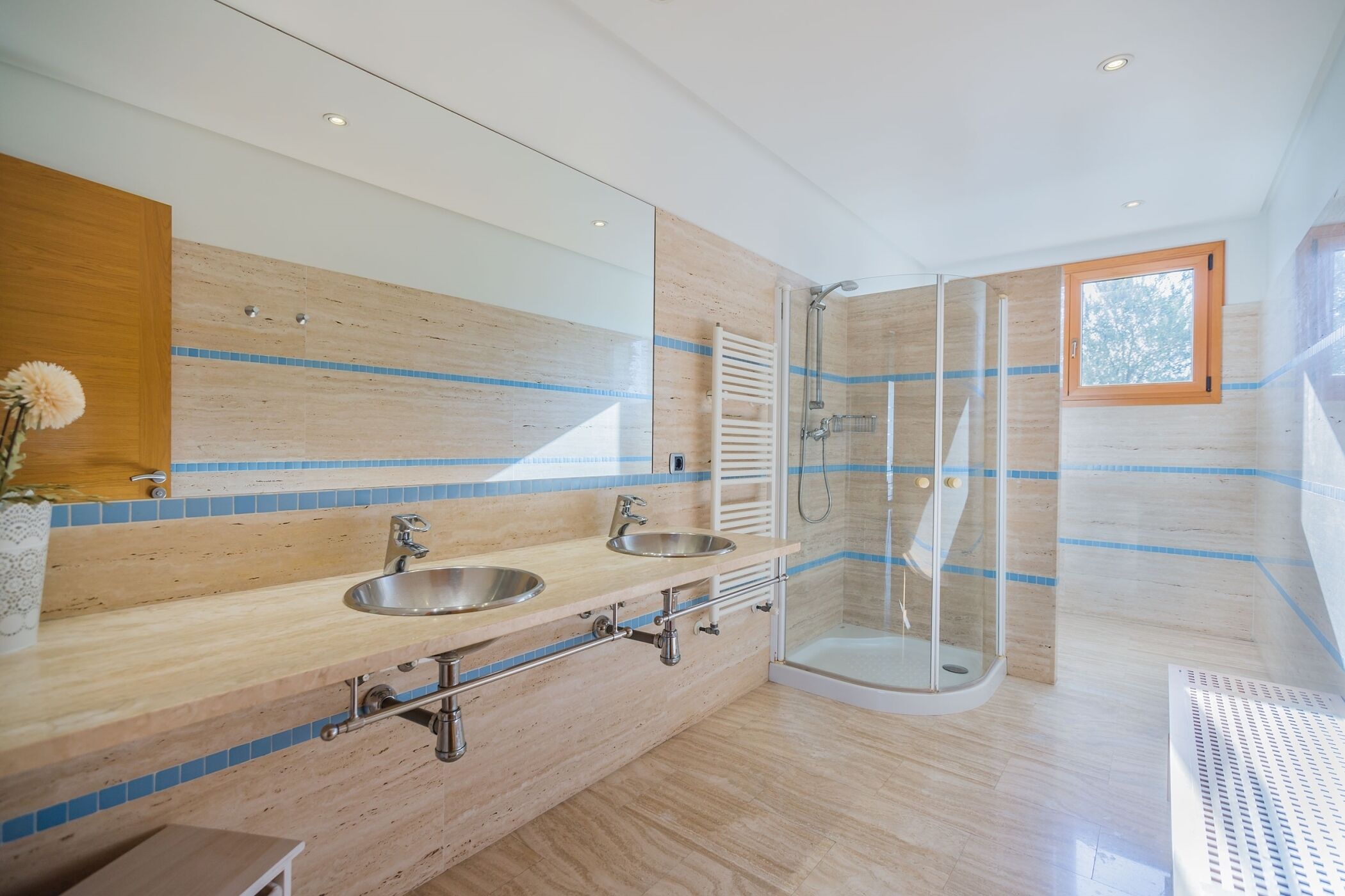 A modern bathroom with a wooden vanity, two sinks, a glass shower enclosure, and a window.