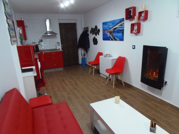 Une cuisine moderne avec des meubles rouges, un plan de travail blanc et une hotte en acier inoxydable. On y trouve un coin repas avec une table blanche et des chaises rouges, ainsi qu’un salon avec un canapé rouge et une cheminée.