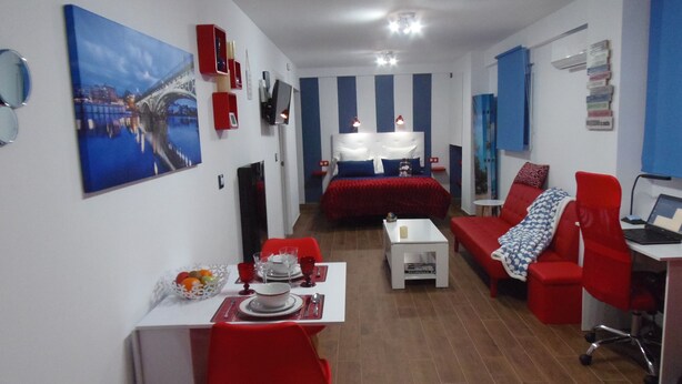 A modern hotel room with a dining area, a bed with a red headboard, a red sofa, and a desk with a laptop. - Beautiful studio in Triana (Sevilla)