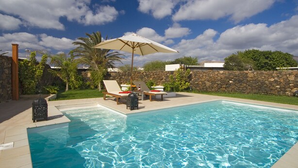 A swimming pool with lounge chairs and umbrellas, surrounded by a stone wall and greenery. - Modern Villa with Heated Pool, Wifi and Air Conditioning in Los Mojones (Puerto del Carmen)