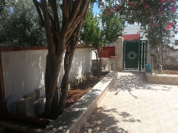 Un chemin pavé mène à une entrée fermée par un portail, ornée d’un motif solaire décoratif, et est bordé d’arbres et d’un petit muret.