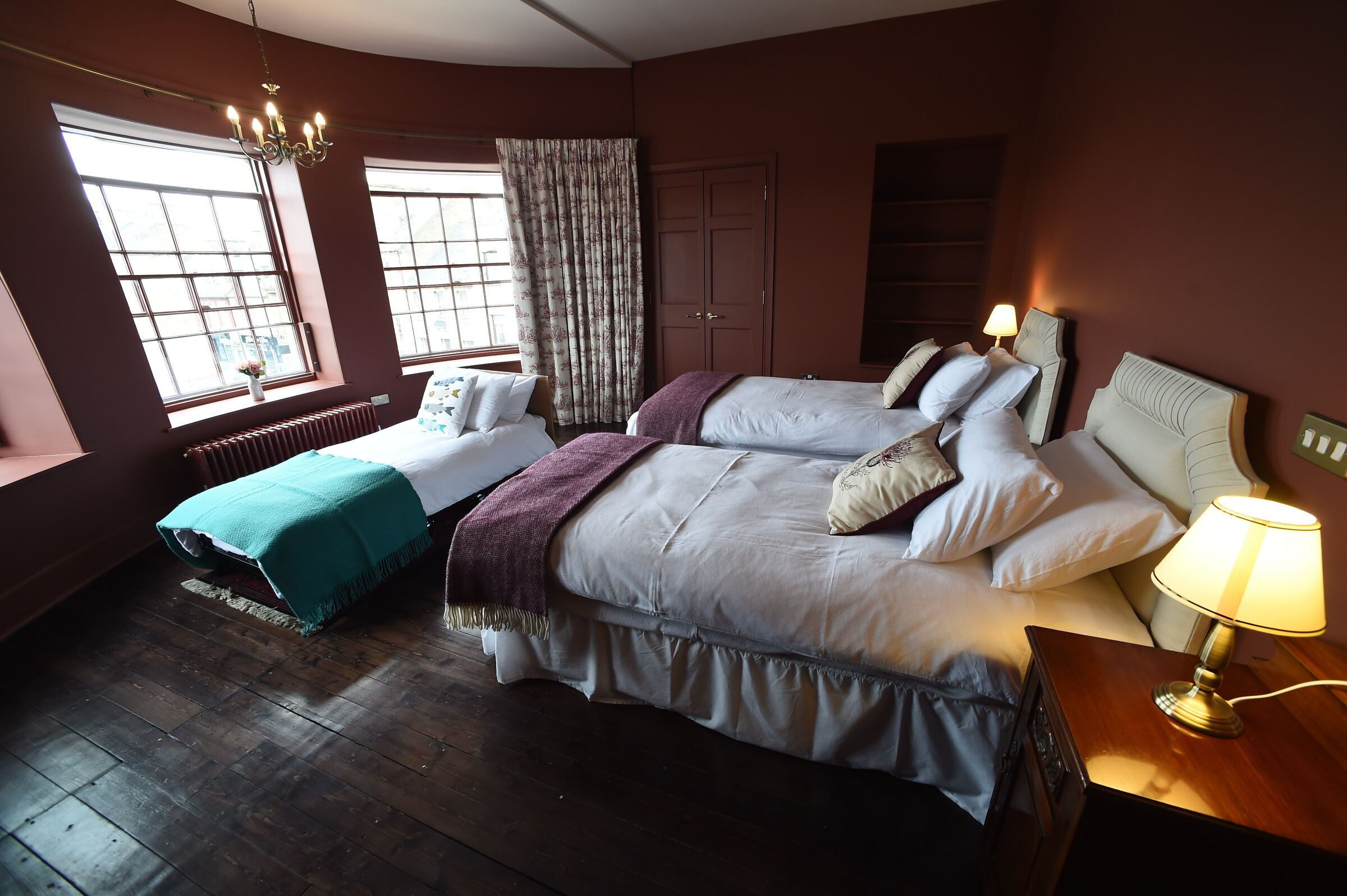 A hotel room with two beds, a window with curtains, a radiator, and a lamp on a bedside table.
