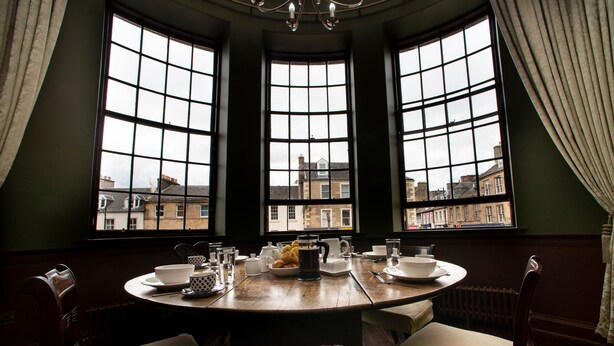 A dining room with a round table set for four, large windows with a view of buildings, and curtains on the windows. - Unique Historic Apartment sleeping 7 close to St Andrews (Cupar)