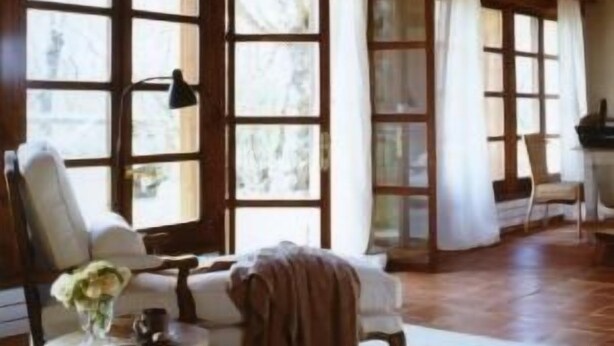 Una habitación amplia con vigas de madera, ventanas grandes y una zona de estar acogedora con una silla y una mesa.