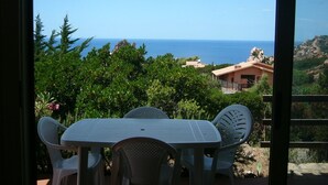 A balcony with a table and chairs, overlooking a coastal landscape with greenery and a building in the distance. - VILLA Belvedere AZZURRA (Costa Paradiso)