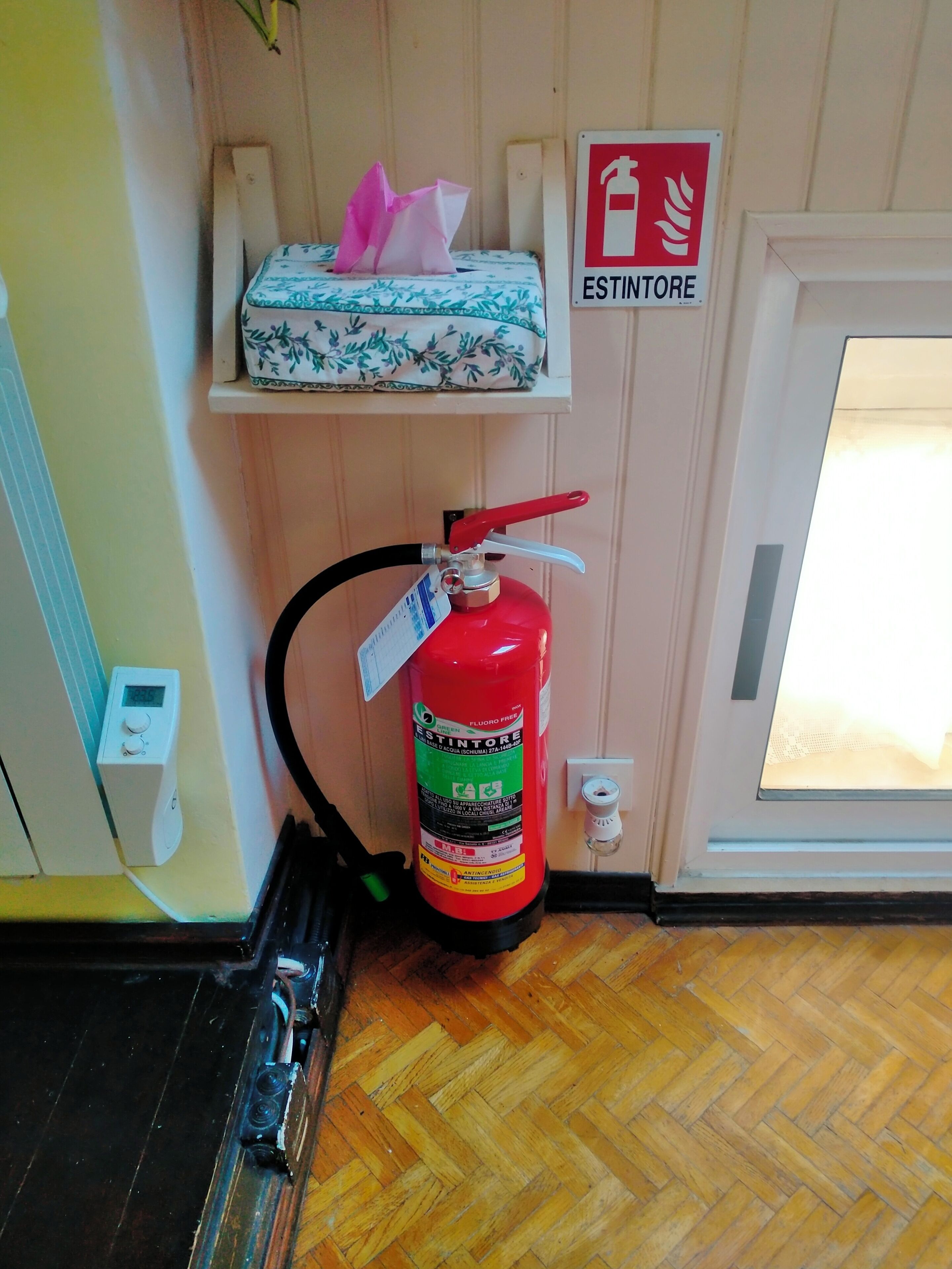 A fire extinguisher mounted on a wall, a digital thermostat, and a sign indicating a fire exit.