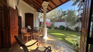 A covered patio area with a wooden table and chairs, a white pillar, and a view of a well-maintained garden with various plants and a fence. - Premium Colonial Villa with Pool, charming Staff + Beach House (Ambalangoda)
