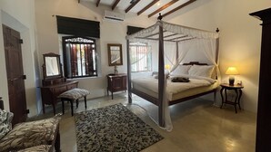 A four-poster canopy bed with a nightstand and lamp, a desk with a chair, a mirror, and a patterned rug in a room with wooden beams and a ceiling fan. - Premium Colonial Villa with Pool, charming Staff + Beach House (Ambalangoda)
